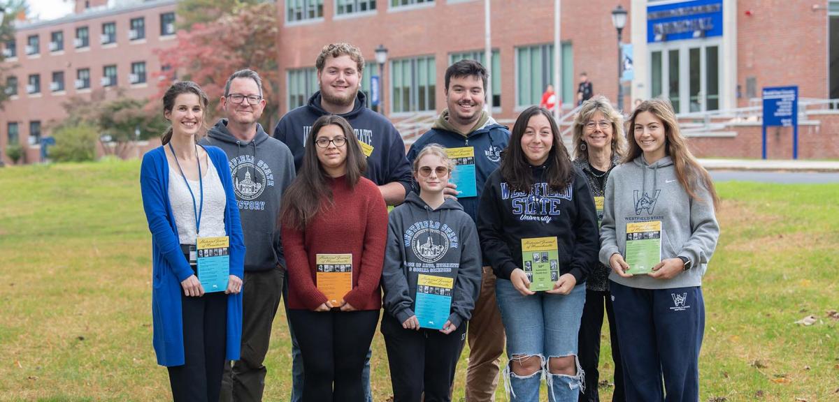 A group of students and professors stand on the Westfield State Campus green holding books.