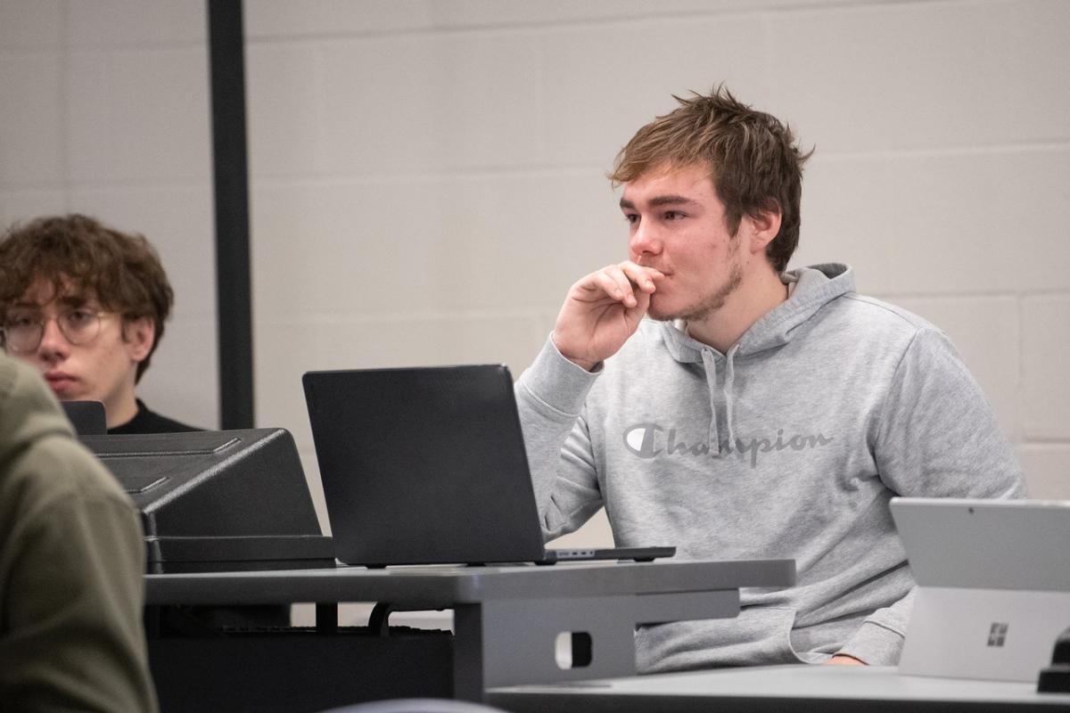 Student in a classroom listening to instructor while working on a laptop.