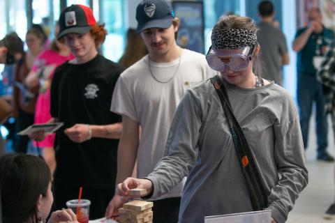 A student with goggles plays Jenga with another student behind him smiling.