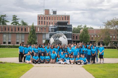 A photo of the Urban Education Summer Bridge Program students wearing a turquoise-colored T-shirt and shorts. They pose in front of the campus globe, which features the brick Ely building in the background.