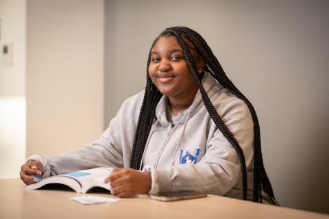 Jakayla Leary, a first-year at the University, sits at a desk with a book open before her. Her long brown hair is down, and she wears a grey sweatshirt with a blue Nestor logo on the front.