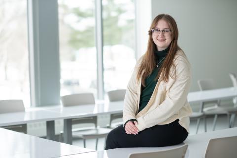 Nicole Dolat, class of 2024. She is dressed in a green shirt, a white over-sweater, black glasses, black pants, and sits on top of a white, glossy desk. The walls behind her are white, and she is in a classroom. Windows along it are open, and there is greenery blurred in the background.