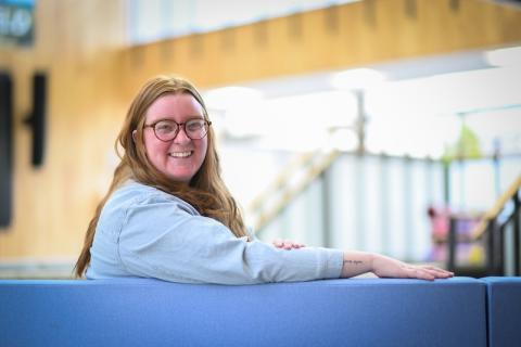 Averie Bye-Dickinson, a graduate student of the Master go Public Administration program. She is sitting in the lobby in Horace Mann, and is wearing a gray, long-sleeve shirt. She has red hair and glasses, and has one arm over the back of the chair she sits on as she smiles at the camera. White light from the background streams in behind her.