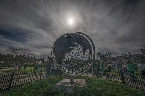 The 2024 total solar eclipse. The silver campus globe is in the middle of the photo, with the sun directly above it. Clouds cover the eclipse, but the lighting is dark and students beyond the globe gather to witness the moon.