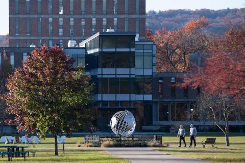 A campus stock photo of the globe and Ely in mid-fall. Red and green trees are on either side of the globe, and two figures are walking in the distance.