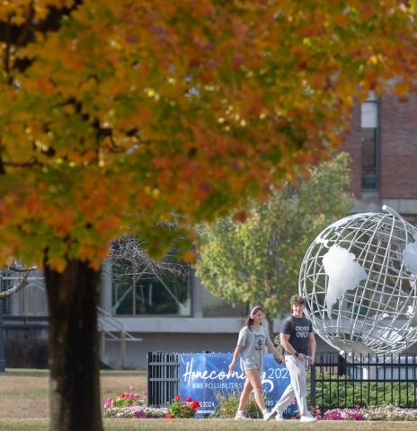 A stock photo of campus, with two students walking by the silver globe near Ely. In the corner of the photo is a tree with green and yellow leaves from autumn.