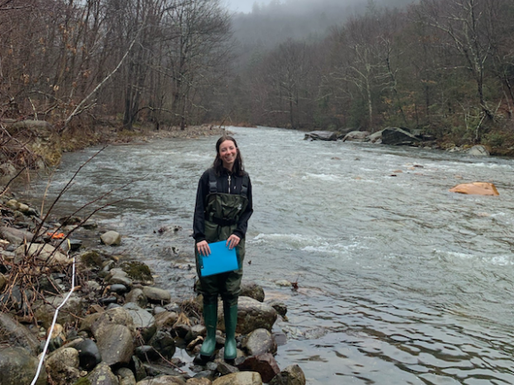 Student Elizabeth Griffin working on her environmental science senior capstone research project on the Westfield River. She stands by the edge and smiles at the camera.
