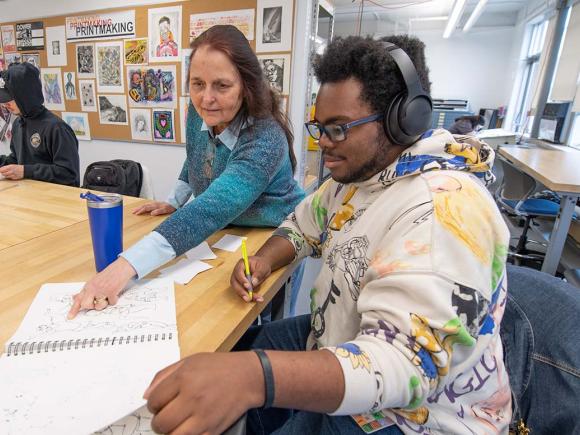 A professor and student review the student’s sketches while seated in an art classroom.