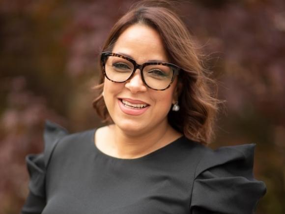 Assistant Director for Student Access and Support, Marjorie Rodriguez. She wears a black shirt and glasses and stands in front of blurred foliage.