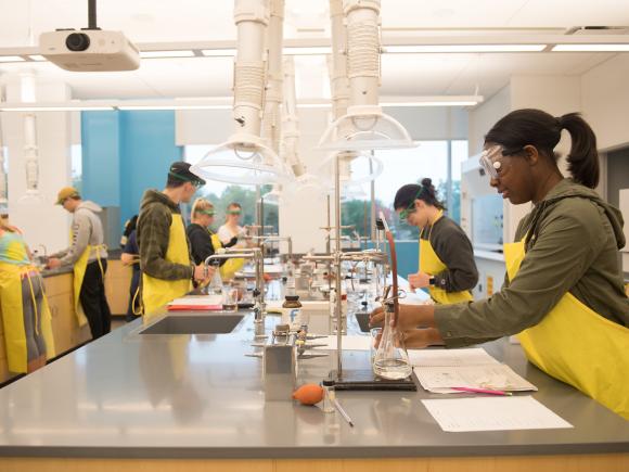 Chemistry lab with students learning hands on wearing lab aprons and eye protection.