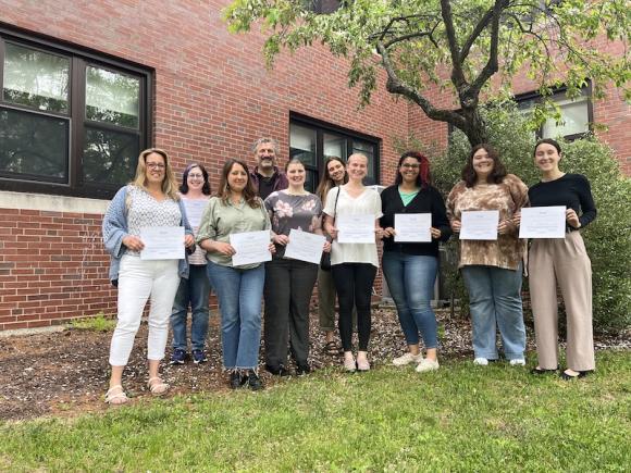 Newly graduated students from the Masters of Social Work program. They're standing with their certificates beside a tree and a brick building on campus.