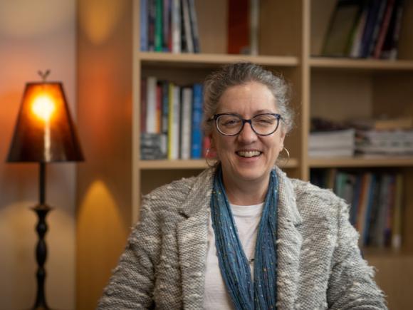 Dr. Kim Tobin, Professor of the Criminal Justice Department. She is sitting in her office and is seated before a bookcase. She wears a gray jacket and blue scarf.