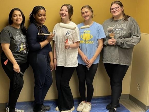 The five students who created a new pediatric waiting room at the MiraVista Behavioral Health Center in Holyoke, MA. Four young women pose in front of a yellow wall, and they're holding paint brushes to show some of the work they've done to create the room.