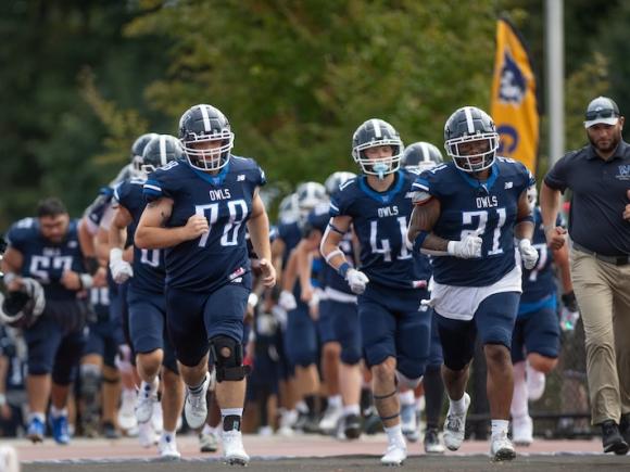 The Westfield State Owl Football team on the 2024 Family Weekend football  game. Lines of students in their dark blue uniforms jog out after being introduced.