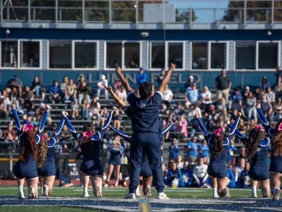 The University's cheerleading team performing in front of a crowd for this year's Homecoming weekend. Owls in blue uniforms face the crowd on bleachers and raise their hands in the air.