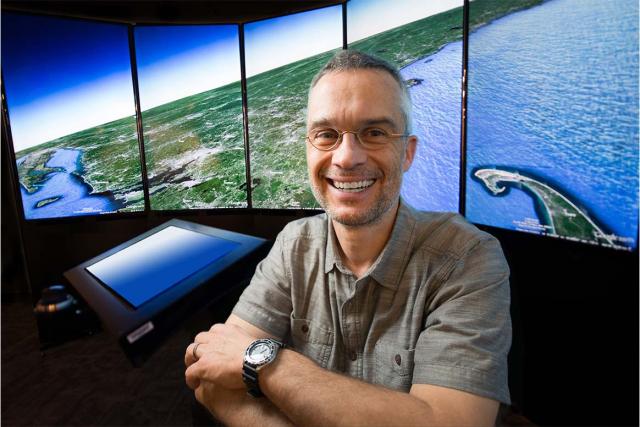 A Westfield State University professor smiles while sitting in front of multiple large screens displaying an aerial view of a coastline.