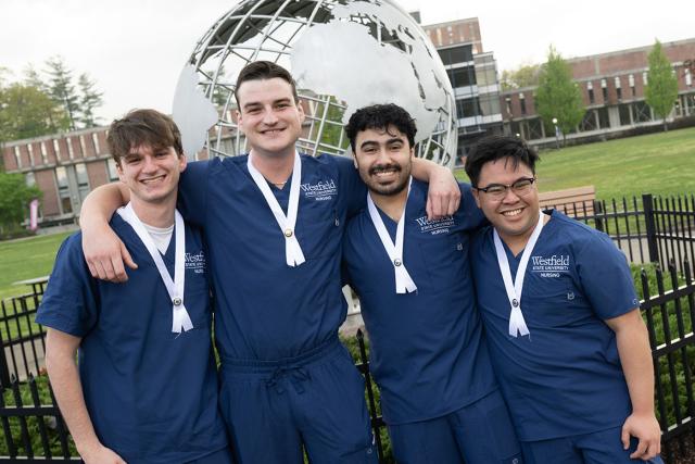 Four nursing students wearing blue scrubs pose for a photo in front of Westfield State University’s globe statue.