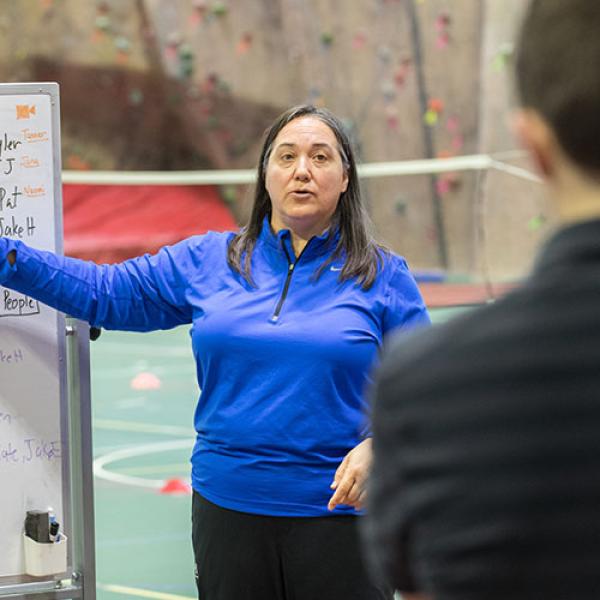 Heidi Bohler, Chair of the Sports Medicine and Human Performance Department at Westfield State University stands on an athletic court in front of a climbing wall while instructing students.