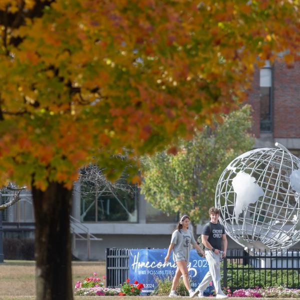 A stock photo of campus, with two students walking by the silver globe near Ely. In the corner of the photo is a tree with green and yellow leaves from autumn.