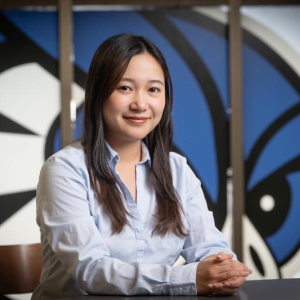 Honatsu Kabu, Class of 2025. A business administration major, Kabu wears a light blue shirt and sits at a desk in front of a Westfield State Owls mural.